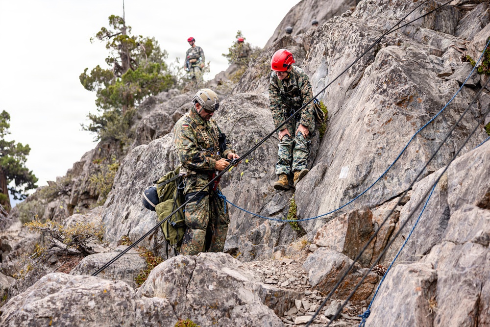 Military personnel in camouflage uniforms are conducting a rock climbing or rappelling training exercise on a rocky cliff face. Multiple individuals are visible, with two in the foreground managing ropes and rigging equipment on the rock face, while two others observe from the top of the ridge. The scene suggests a multinational military training exercise based on the differing camouflage patterns worn by participants.