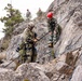 Military personnel in camouflage uniforms are conducting a rock climbing or rappelling training exercise on a rocky cliff face. Multiple individuals are visible, with two in the foreground managing ropes and rigging equipment on the rock face, while two others observe from the top of the ridge. The scene suggests a multinational military training exercise based on the differing camouflage patterns worn by participants.