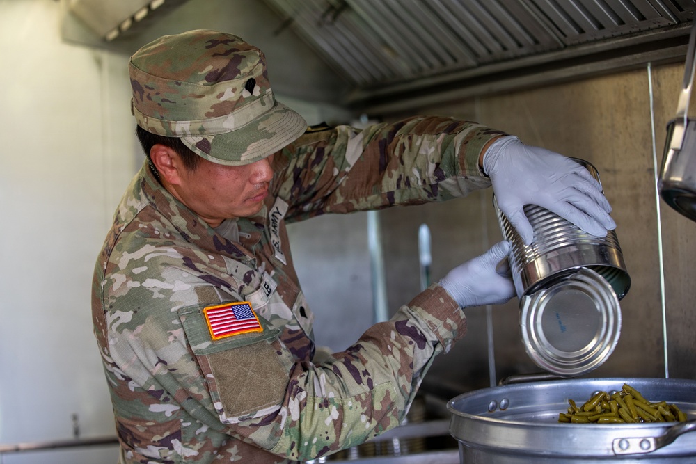 A U.S. Army soldier in OCP (Operational Camouflage Pattern) uniform is preparing food in a field kitchen environment. The soldier is wearing latex gloves and pouring the contents of a large metal can into a cooking vessel. Green beans are visible in the receiving container, indicating food service or field mess operations.