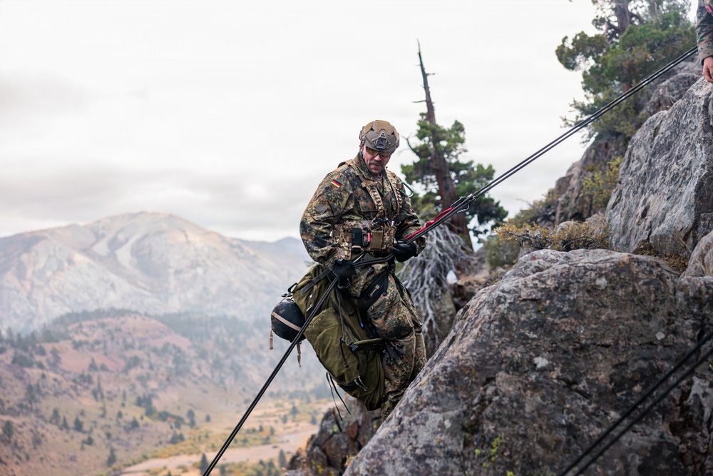 A military soldier in camouflage uniform is rappelling or climbing down a rocky cliff face in a mountainous environment. The soldier is equipped with full tactical gear including a helmet, harness, large backpack, and is managing ropes on the rock face. The background reveals a dramatic mountain landscape under overcast skies.
