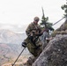 A military soldier in camouflage uniform is rappelling or climbing down a rocky cliff face in a mountainous environment. The soldier is equipped with full tactical gear including a helmet, harness, large backpack, and is managing ropes on the rock face. The background reveals a dramatic mountain landscape under overcast skies.