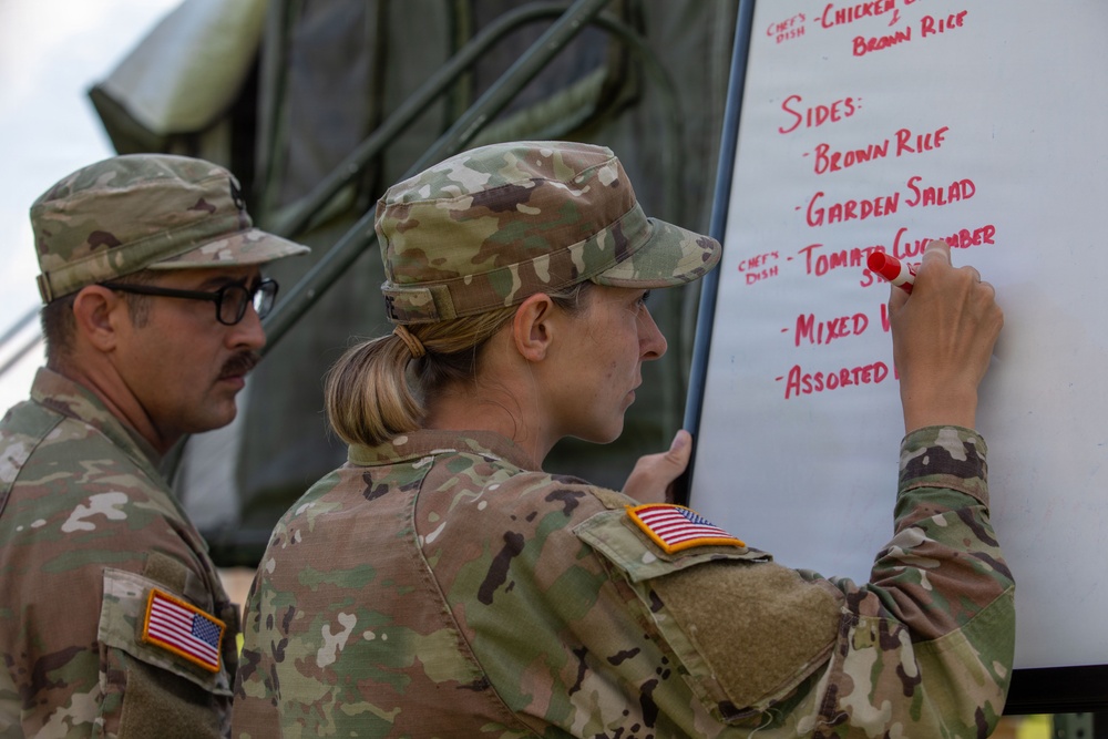 Two U.S. Army soldiers in OCP (Operational Camouflage Pattern) uniforms are writing a menu on a whiteboard outdoors near a military tent. A female soldier is actively writing food items in red marker while a male soldier with glasses observes. The whiteboard displays a meal menu including sides such as Brown Rice, Garden Salad, Tomato Cucumber, Mixed Vegetables, and Assorted items.
