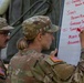 Two U.S. Army soldiers in OCP (Operational Camouflage Pattern) uniforms are writing a menu on a whiteboard outdoors near a military tent. A female soldier is actively writing food items in red marker while a male soldier with glasses observes. The whiteboard displays a meal menu including sides such as Brown Rice, Garden Salad, Tomato Cucumber, Mixed Vegetables, and Assorted items.