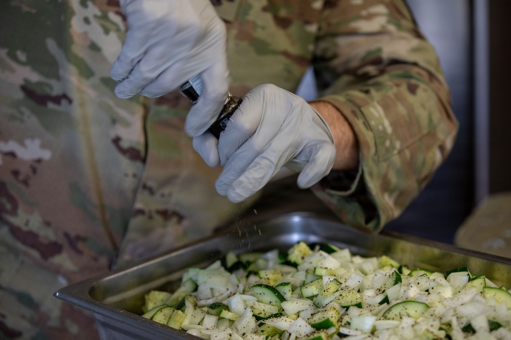 A person wearing military camouflage (OCP/MultiCam pattern) uniform and white latex gloves is seasoning a large metal hotel pan filled with chopped vegetables. The vegetables appear to be zucchini and onions, and seasoning (likely pepper or a spice blend) is visibly falling from a small container onto the food. This appears to be a military food service or field kitchen preparation scenario.