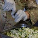 A person wearing military camouflage (OCP/MultiCam pattern) uniform and white latex gloves is seasoning a large metal hotel pan filled with chopped vegetables. The vegetables appear to be zucchini and onions, and seasoning (likely pepper or a spice blend) is visibly falling from a small container onto the food. This appears to be a military food service or field kitchen preparation scenario.