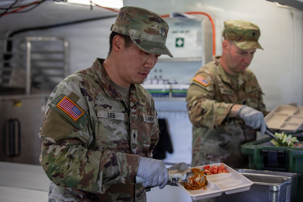 Two U.S. Army soldiers in Operational Camouflage Pattern (OCP) uniforms are serving food in what appears to be a military field kitchen or dining facility. The foreground soldier, identified by the name tape 'LEE' and 'U.S. ARMY' tape, is plating food onto a compartmentalized tray while wearing food-service gloves. A second soldier in the background is also handling food items from a green container.