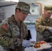 Two U.S. Army soldiers in Operational Camouflage Pattern (OCP) uniforms are serving food in what appears to be a military field kitchen or dining facility. The foreground soldier, identified by the name tape 'LEE' and 'U.S. ARMY' tape, is plating food onto a compartmentalized tray while wearing food-service gloves. A second soldier in the background is also handling food items from a green container.