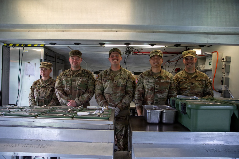 Five U.S. Army soldiers in OCP (Operational Camouflage Pattern) uniforms are posing together behind a military field kitchen serving line. They appear to be food service personnel (cooks/92G MOS) staffing a mobile kitchen trailer (MKT). The soldiers are smiling for what appears to be an official or informal group photo.