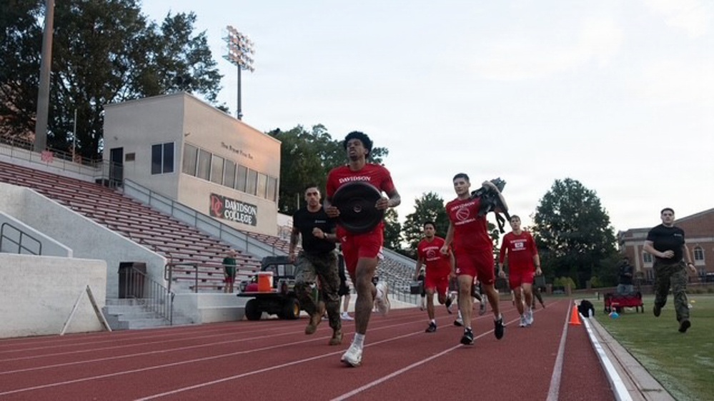 A group of athletes and military personnel are running on a red track at Davidson College stadium. The lead runner, wearing a red Davidson jersey, is carrying a weight plate while others follow behind. The scene appears to be a combined athletic and military fitness training exercise.