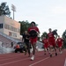 A group of athletes and military personnel are running on a red track at Davidson College stadium. The lead runner, wearing a red Davidson jersey, is carrying a weight plate while others follow behind. The scene appears to be a combined athletic and military fitness training exercise.