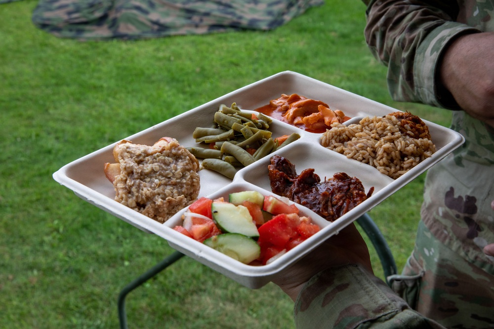 A person wearing camouflage military uniform holds a compartmentalized food tray containing multiple dishes, likely a field meal served outdoors. The tray is a disposable multi-section container with six compartments filled with various foods. The setting appears to be a grassy outdoor area, consistent with a military field dining environment.