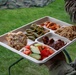 A person wearing camouflage military uniform holds a compartmentalized food tray containing multiple dishes, likely a field meal served outdoors. The tray is a disposable multi-section container with six compartments filled with various foods. The setting appears to be a grassy outdoor area, consistent with a military field dining environment.