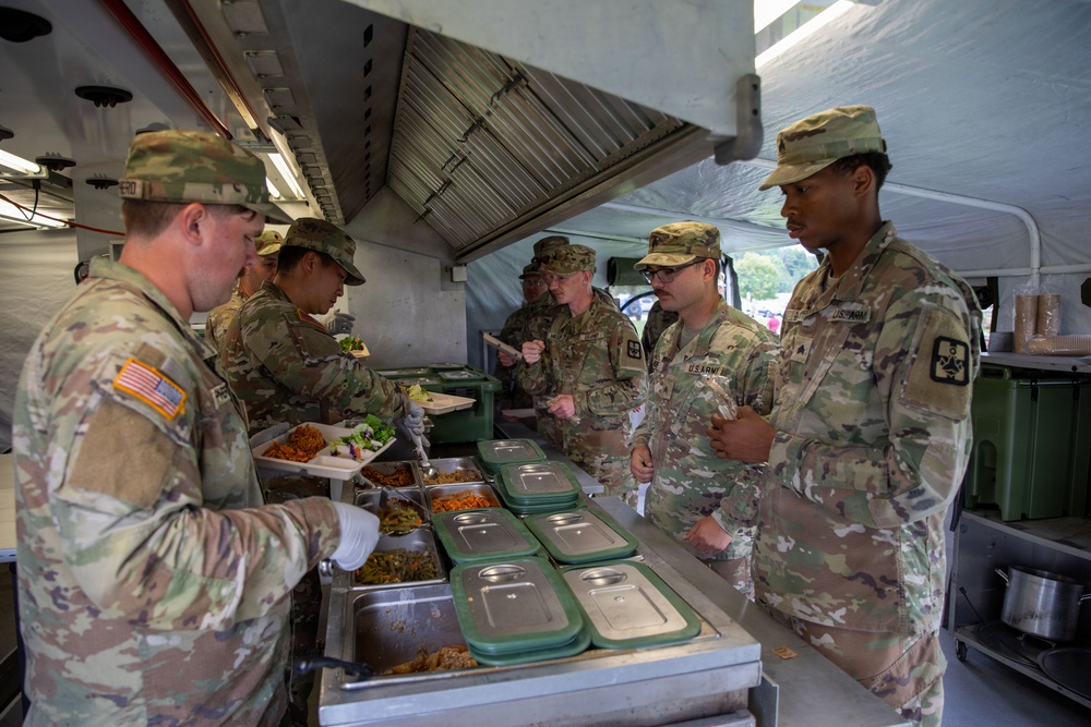 U.S. Army soldiers are being served food at a field kitchen or mobile dining facility. A soldier wearing white food-service gloves is serving meals from stainless steel food containers to a line of uniformed personnel. The setting appears to be a temporary or field mess operation under a canopy structure.