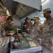 U.S. Army soldiers are being served food at a field kitchen or mobile dining facility. A soldier wearing white food-service gloves is serving meals from stainless steel food containers to a line of uniformed personnel. The setting appears to be a temporary or field mess operation under a canopy structure.