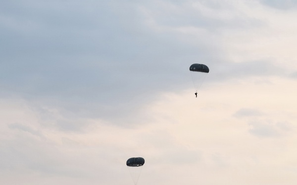 A military airborne operation is underway, showing multiple paratroopers descending under dark round parachutes over a flat, open drop zone. At least five parachutists are visible at various altitudes, with some near landing and others still high in the sky. A military vehicle is visible on the ground at the left edge of the landing zone, surrounded by sparse treeline in the background.