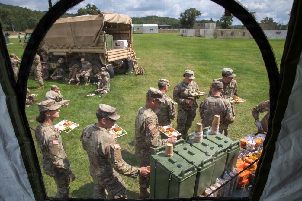The image shows U.S. Army soldiers in camouflage uniforms receiving a field meal during what appears to be a training exercise or field operation. The photo is taken from inside a tent or shelter, looking outward at soldiers holding food trays and gathering around green insulated food containers. Additional soldiers are seated on the grass near a military truck in the background.