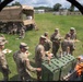 The image shows U.S. Army soldiers in camouflage uniforms receiving a field meal during what appears to be a training exercise or field operation. The photo is taken from inside a tent or shelter, looking outward at soldiers holding food trays and gathering around green insulated food containers. Additional soldiers are seated on the grass near a military truck in the background.