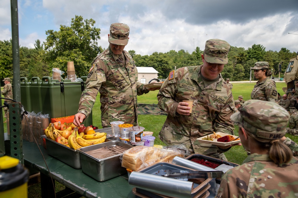 U.S. Army soldiers are gathered at an outdoor field mess station, serving and receiving food during what appears to be a training exercise or field operation. Two soldiers in the foreground, identified by name tapes as 'HUDDLESTON' and 'BASNA' (partially visible), are actively engaged at the food service table. Multiple soldiers in OCP (Operational Camouflage Pattern) uniforms are visible in the background near military vehicles.