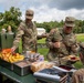U.S. Army soldiers are gathered at an outdoor field mess station, serving and receiving food during what appears to be a training exercise or field operation. Two soldiers in the foreground, identified by name tapes as 'HUDDLESTON' and 'BASNA' (partially visible), are actively engaged at the food service table. Multiple soldiers in OCP (Operational Camouflage Pattern) uniforms are visible in the background near military vehicles.