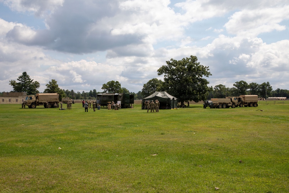 A military field exercise or training operation is taking place on an open grassy area. Several tan/beige military tactical trucks are positioned across the field alongside green military tents or shelters. Personnel in military uniforms are gathered around the equipment and structures.