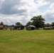 A military field exercise or training operation is taking place on an open grassy area. Several tan/beige military tactical trucks are positioned across the field alongside green military tents or shelters. Personnel in military uniforms are gathered around the equipment and structures.