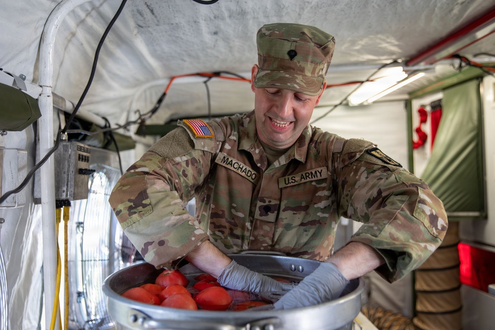 A U.S. Army soldier named Machado is smiling while handling sliced tomatoes in a large metal bowl inside what appears to be a military field kitchen tent. The soldier is wearing full OCP (Operational Camouflage Pattern) uniform with an American flag patch and blue/gray food-service gloves. The setting suggests a forward operating base or field exercise food preparation environment.