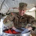 A U.S. Army soldier named Machado is smiling while handling sliced tomatoes in a large metal bowl inside what appears to be a military field kitchen tent. The soldier is wearing full OCP (Operational Camouflage Pattern) uniform with an American flag patch and blue/gray food-service gloves. The setting suggests a forward operating base or field exercise food preparation environment.