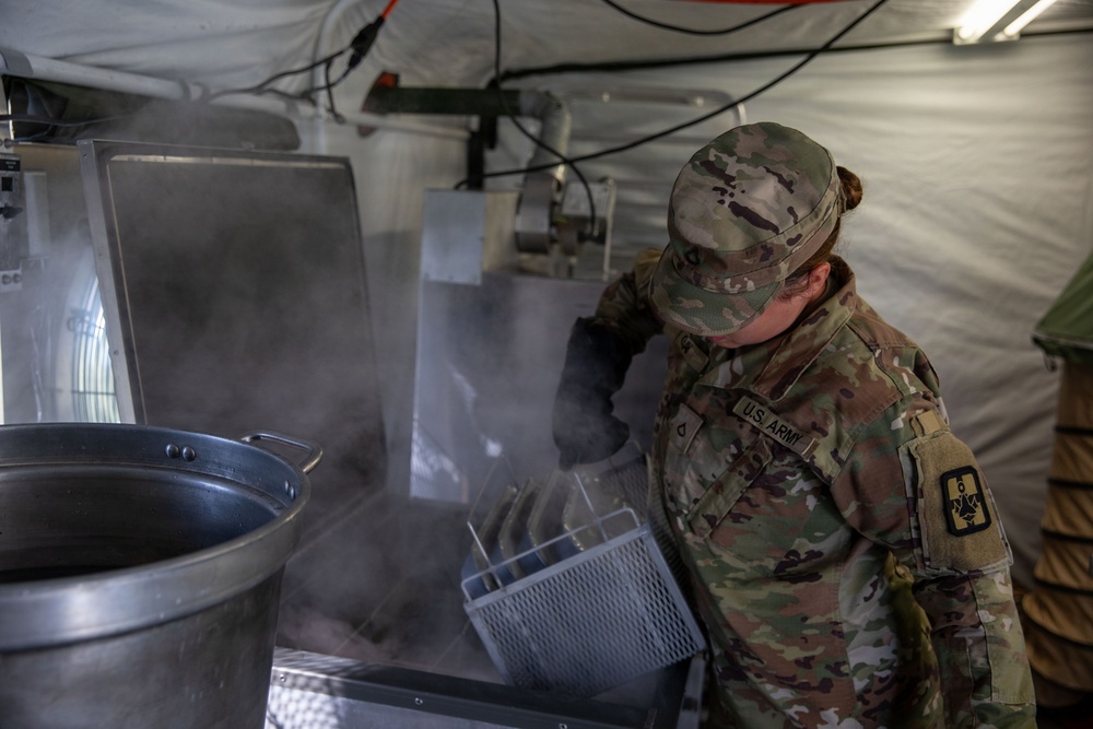 A U.S. Army soldier in Operational Camouflage Pattern (OCP) uniform is working in a field kitchen environment, handling what appears to be a steaming dishwashing or food preparation station inside a military tent. Steam is visibly rising from the equipment, suggesting hot water or cooking operations are underway. The setting appears to be a temporary military field kitchen or mess facility.