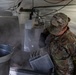 A U.S. Army soldier in Operational Camouflage Pattern (OCP) uniform is working in a field kitchen environment, handling what appears to be a steaming dishwashing or food preparation station inside a military tent. Steam is visibly rising from the equipment, suggesting hot water or cooking operations are underway. The setting appears to be a temporary military field kitchen or mess facility.