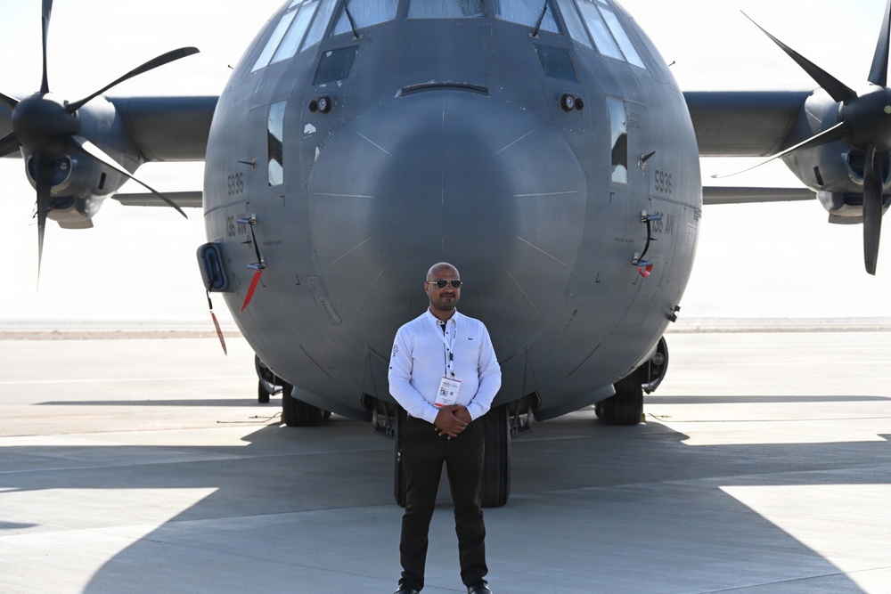 A man wearing a white shirt, black pants, and sunglasses stands in front of a large military turboprop aircraft on a tarmac. He is wearing a lanyard with an identification badge, suggesting attendance at an airshow or official aviation event. The aircraft behind him is a gray military transport plane, identifiable as a C-130 Hercules variant, with visible serial numbers on the fuselage.