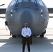 A man wearing a white shirt, black pants, and sunglasses stands in front of a large military turboprop aircraft on a tarmac. He is wearing a lanyard with an identification badge, suggesting attendance at an airshow or official aviation event. The aircraft behind him is a gray military transport plane, identifiable as a C-130 Hercules variant, with visible serial numbers on the fuselage.