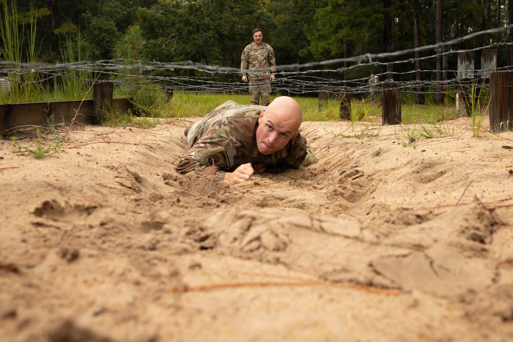 A bald soldier in camouflage uniform is low-crawling through sandy terrain beneath strands of barbed wire as part of a military obstacle course training exercise. A second soldier in camouflage stands upright in the background, observing or waiting their turn. The setting appears to be a wooded military training facility with wooden post barriers supporting the wire obstacle.