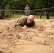 A bald soldier in camouflage uniform is low-crawling through sandy terrain beneath strands of barbed wire as part of a military obstacle course training exercise. A second soldier in camouflage stands upright in the background, observing or waiting their turn. The setting appears to be a wooded military training facility with wooden post barriers supporting the wire obstacle.