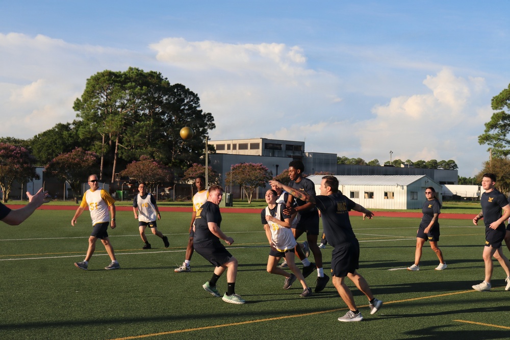 A group of people are playing an outdoor ball sport on a green artificial turf field. Two teams are distinguishable by their uniforms — one wearing white jerseys and the other wearing dark (black/navy) shirts with gold lettering. A gold/yellow ball is visible in the air above the players during active gameplay.
