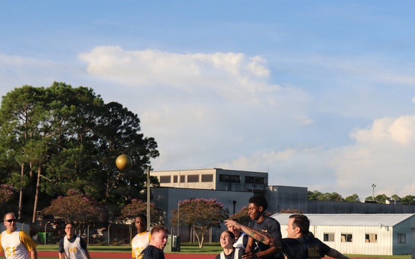 A group of people are playing an outdoor ball sport on a green artificial turf field. Two teams are distinguishable by their uniforms — one wearing white jerseys and the other wearing dark (black/navy) shirts with gold lettering. A gold/yellow ball is visible in the air above the players during active gameplay.