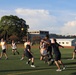 A group of people are playing an outdoor ball sport on a green artificial turf field. Two teams are distinguishable by their uniforms — one wearing white jerseys and the other wearing dark (black/navy) shirts with gold lettering. A gold/yellow ball is visible in the air above the players during active gameplay.