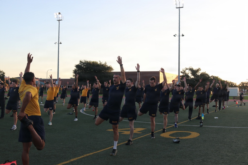 A large group of military personnel, predominantly wearing dark navy blue shorts and shirts with 'NAVY' markings, are performing synchronized physical training exercises on an athletic field. They appear to be doing a balance or stretching exercise with arms raised and one knee lifted. The session is led by an instructor in a yellow shirt on the left side of the frame, and takes place during early morning or evening hours based on the lighting.