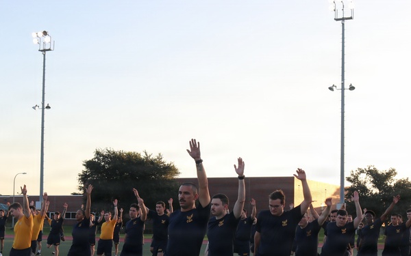 A large group of military personnel, predominantly wearing dark navy blue shorts and shirts with 'NAVY' markings, are performing synchronized physical training exercises on an athletic field. They appear to be doing a balance or stretching exercise with arms raised and one knee lifted. The session is led by an instructor in a yellow shirt on the left side of the frame, and takes place during early morning or evening hours based on the lighting.