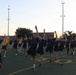 A large group of military personnel, predominantly wearing dark navy blue shorts and shirts with 'NAVY' markings, are performing synchronized physical training exercises on an athletic field. They appear to be doing a balance or stretching exercise with arms raised and one knee lifted. The session is led by an instructor in a yellow shirt on the left side of the frame, and takes place during early morning or evening hours based on the lighting.
