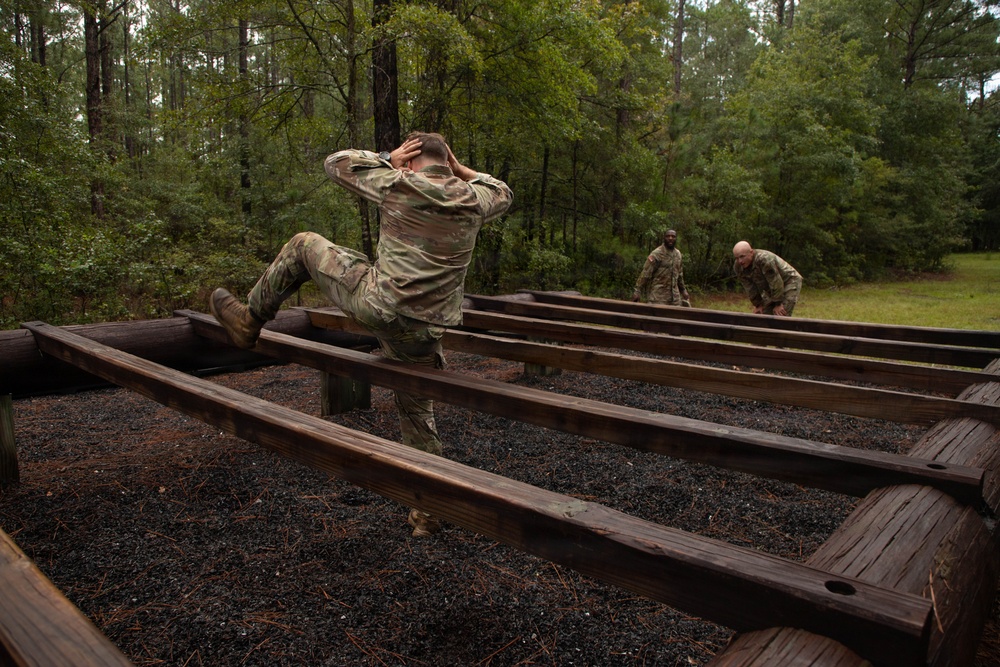 Military personnel in OCP (Operational Camouflage Pattern) uniforms are navigating a wooden obstacle course in a forested training area. The foreground soldier is performing a low crawl or hurdle maneuver over parallel wooden beams with hands behind their head, while two additional soldiers approach in the background. The setting appears to be a military physical training or obstacle course facility with dark gravel ground cover.