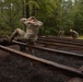 Military personnel in OCP (Operational Camouflage Pattern) uniforms are navigating a wooden obstacle course in a forested training area. The foreground soldier is performing a low crawl or hurdle maneuver over parallel wooden beams with hands behind their head, while two additional soldiers approach in the background. The setting appears to be a military physical training or obstacle course facility with dark gravel ground cover.