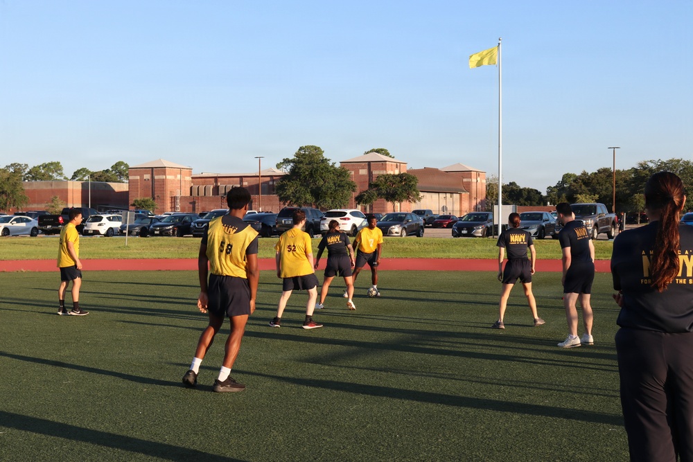 A group of individuals wearing U.S. Navy-branded yellow and dark athletic uniforms are playing soccer or participating in a physical training activity on a green turf field. Several players wear numbered jerseys (58, 52 visible) and Navy-branded shirts. The setting appears to be a military installation, evidenced by the yellow flag on a flagpole and brick institutional buildings in the background.
