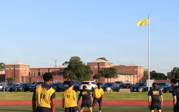 A group of individuals wearing U.S. Navy-branded yellow and dark athletic uniforms are playing soccer or participating in a physical training activity on a green turf field. Several players wear numbered jerseys (58, 52 visible) and Navy-branded shirts. The setting appears to be a military installation, evidenced by the yellow flag on a flagpole and brick institutional buildings in the background.