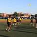 A group of individuals wearing U.S. Navy-branded yellow and dark athletic uniforms are playing soccer or participating in a physical training activity on a green turf field. Several players wear numbered jerseys (58, 52 visible) and Navy-branded shirts. The setting appears to be a military installation, evidenced by the yellow flag on a flagpole and brick institutional buildings in the background.