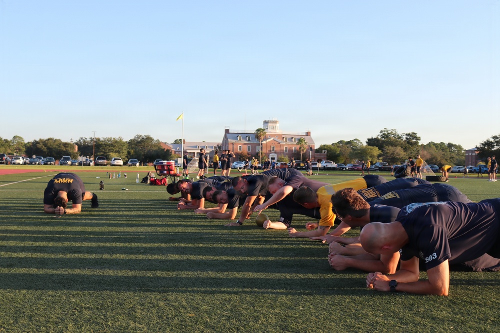 A group of individuals wearing dark Navy-branded athletic shirts are performing a plank or push-up exercise in a line on a green turf field. The scene appears to be a military physical training session, likely U.S. Navy, taking place outdoors during early morning or late afternoon based on the low-angle sunlight. A large brick institutional building is visible in the background along with additional participants and parked vehicles.