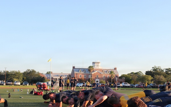 A group of individuals wearing dark Navy-branded athletic shirts are performing a plank or push-up exercise in a line on a green turf field. The scene appears to be a military physical training session, likely U.S. Navy, taking place outdoors during early morning or late afternoon based on the low-angle sunlight. A large brick institutional building is visible in the background along with additional participants and parked vehicles.