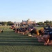 A group of individuals wearing dark Navy-branded athletic shirts are performing a plank or push-up exercise in a line on a green turf field. The scene appears to be a military physical training session, likely U.S. Navy, taking place outdoors during early morning or late afternoon based on the low-angle sunlight. A large brick institutional building is visible in the background along with additional participants and parked vehicles.