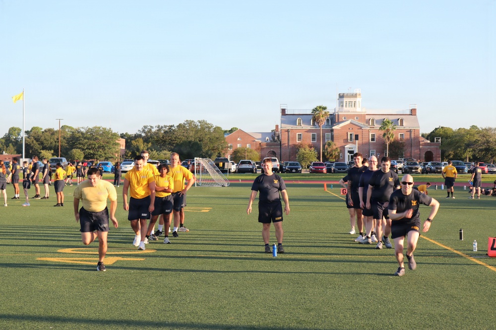 A group of individuals wearing U.S. Navy physical training uniforms (yellow and dark navy/black shirts with 'NAVY' shorts) are conducting physical fitness training on a green athletic field. The activity appears to be a military physical readiness test or group PT session, with participants running and walking across the turf. A large historic red-brick building is visible in the background, consistent with a military installation or naval station.