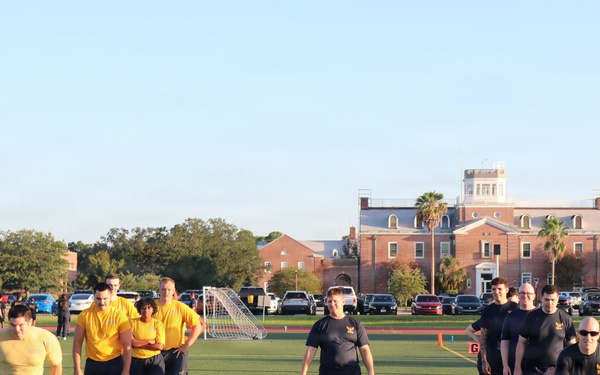 A group of individuals wearing U.S. Navy physical training uniforms (yellow and dark navy/black shirts with 'NAVY' shorts) are conducting physical fitness training on a green athletic field. The activity appears to be a military physical readiness test or group PT session, with participants running and walking across the turf. A large historic red-brick building is visible in the background, consistent with a military installation or naval station.