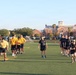 A group of individuals wearing U.S. Navy physical training uniforms (yellow and dark navy/black shirts with 'NAVY' shorts) are conducting physical fitness training on a green athletic field. The activity appears to be a military physical readiness test or group PT session, with participants running and walking across the turf. A large historic red-brick building is visible in the background, consistent with a military installation or naval station.